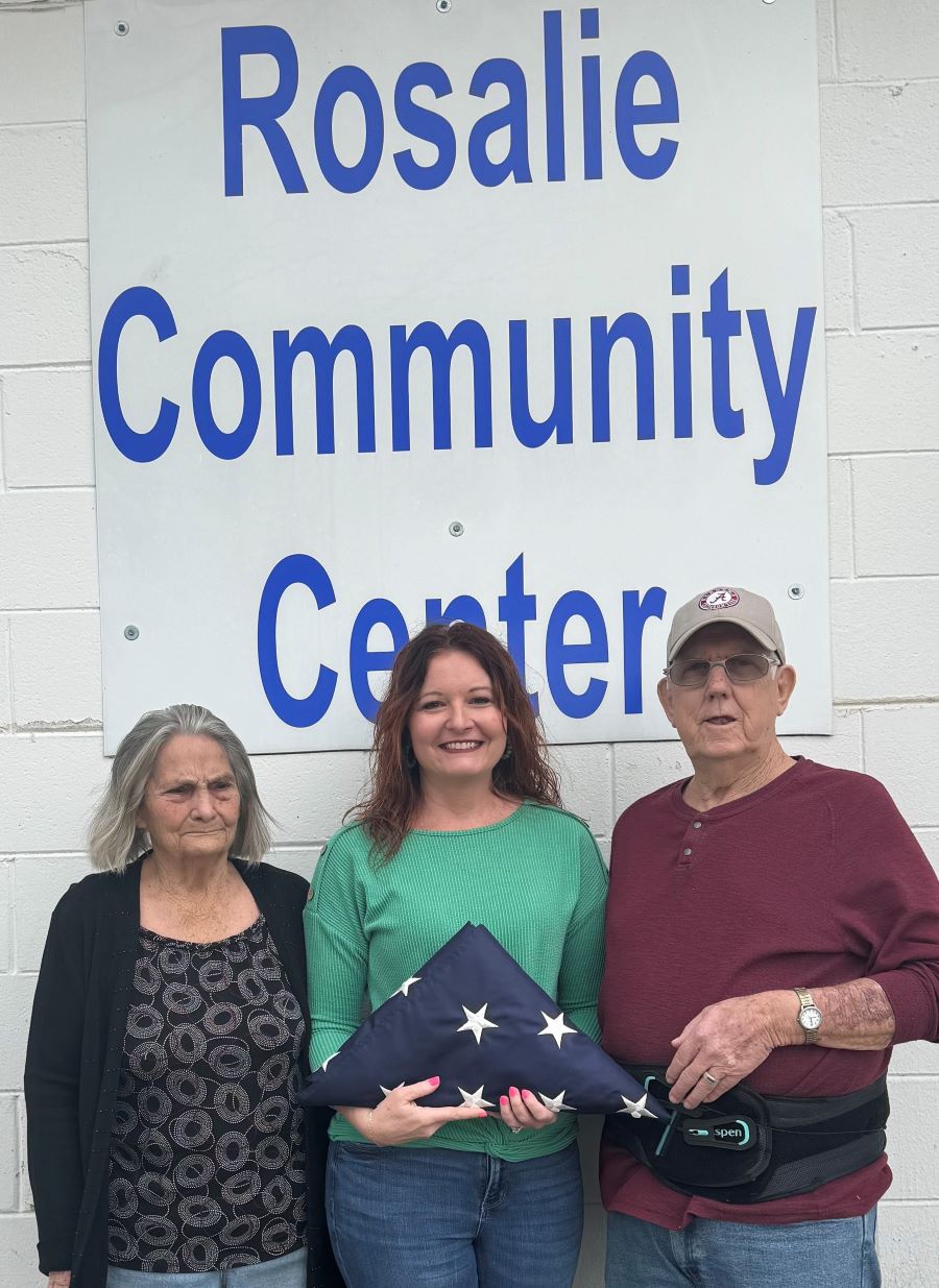 Three people stand together. The woman in the middle is holding a folded American flag. They are standing in front of a sign that says "Rosalie Community Center."