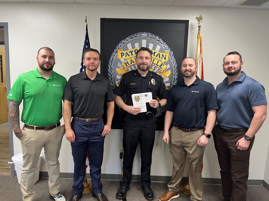 Five men stand together for a photo. The center person is wearing a police uniform. On the wall behind them is a seal for the Hartselle Police Department.