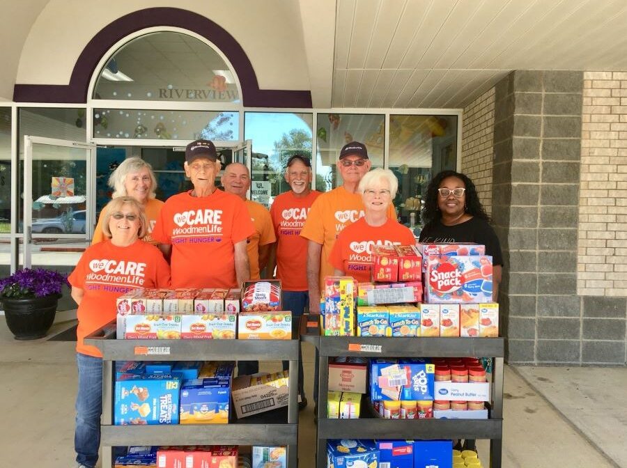 Eight people stand together for the photo. They are standing around two carts full of nonperishable food, including canned fruit, peanut butter and mac & cheese. Seven of the people are wearing orange WoodmenLife t-shirts that say "WeCare WoodmenLife Fight Hunger."