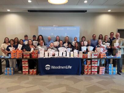 A group of 26 people pose for a photo. They are standing behind a long table that's filled with shelf-stable food items, which will be assembled into the breakfast bags. Several people are holding up white paper bags that are decorated with friendly and inspirational messages. Hanging in front of the table is a navy blue WoodmenLife-branded banner, and behind the group of people is a light blue background with the WoodmenLife logo.