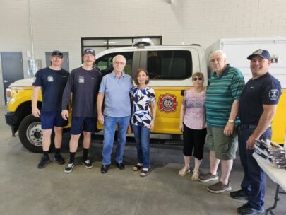 Photo shows seven people standing together in front of a yellow fire station pickup truck. Three of the people in the picture are wearing firefighter T-shirts and hats.