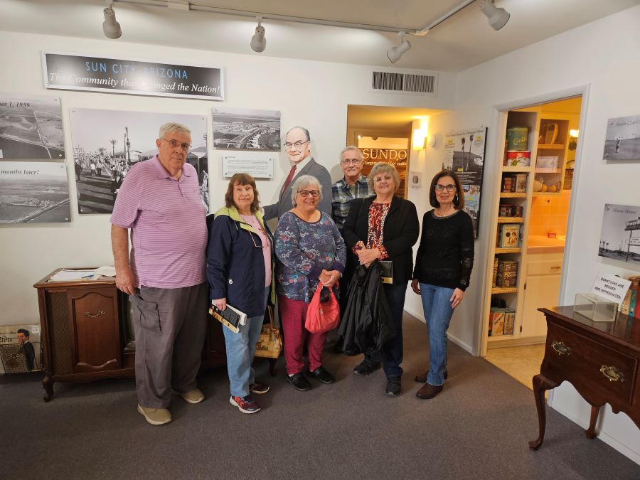 Six people are posing at the Del Webb Sun Cities Museum. Behind them is a life-size cardboard cutout of a man. On the walls are large pictures.