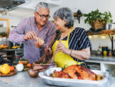 Stock photo shows an older man and woman cooking in the kitchen. The woman is wearing a yellow apron and mixing salad, and the man is grinding pepper over the salad. Also on the counter is a variety of other veggies, as well as a cooked chicken. The couple are looking at each other and smiling.