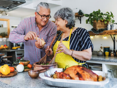 Stock photo shows an older man and woman cooking in the kitchen. The woman is wearing a yellow apron and mixing salad, and the man is grinding pepper over the salad. Also on the counter is a variety of other veggies, as well as a cooked chicken. The couple are looking at each other and smiling.