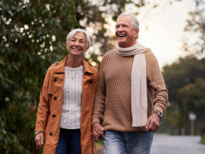 Stock photo shows an older man and woman walking outside. She is wearing a coat, and he is wearing a scarf. Both are smiling.