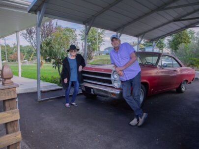 A man and a woman stand on their driveway alongside a classic car. The car is red, and they are both leaning on it. Behind them is green grass and trees.