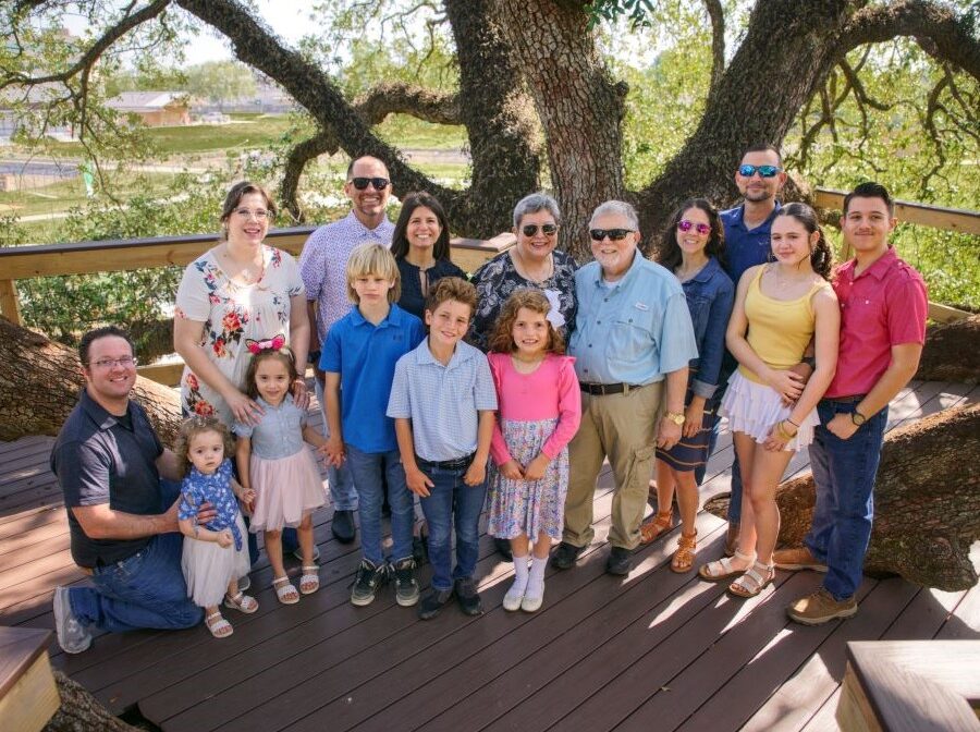 Photo shows a family with 15 people -- it includes two grandparents, their three adult children and their spouses, and seven grandchildren. They are standing outside on a wooden deck, and there is a large tree directly behind them.