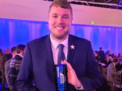 WoodmenLife associate Chad Benner stands holding his Midlands Business Journal 40 Under 40 award. He is wearing a suit, and behind him are people sitting/standing around tables at the awards ceremony.