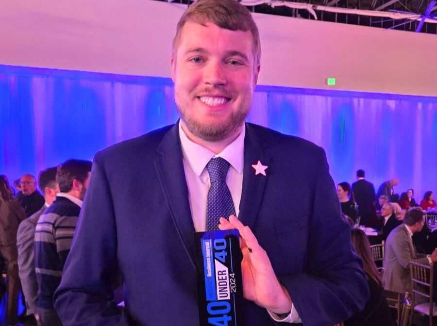 WoodmenLife associate Chad Benner stands holding his Midlands Business Journal 40 Under 40 award. He is wearing a suit, and behind him are people sitting/standing around tables at the awards ceremony.