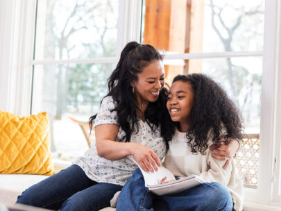 Stock photo shows an adult woman and a girl sitting on a window seat. The woman's arm is around the girl, and the girl is holding a book in her lap. Both are smiling. There are pillows on the seat beside them, and it is bright and sunny outside the window behind them.