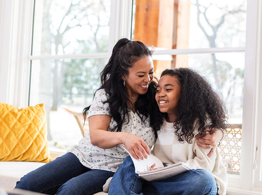 Stock photo shows an adult woman and a girl sitting on a window seat. The woman's arm is around the girl, and the girl is holding a book in her lap. Both are smiling. There are pillows on the seat beside them, and it is bright and sunny outside the window behind them.