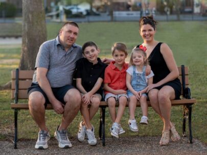 A family of five sits together on an outdoor bench. The dad sits on the left and the mom sits on the right; between them are two boys and a girl. Behind them is green grass and a tree.