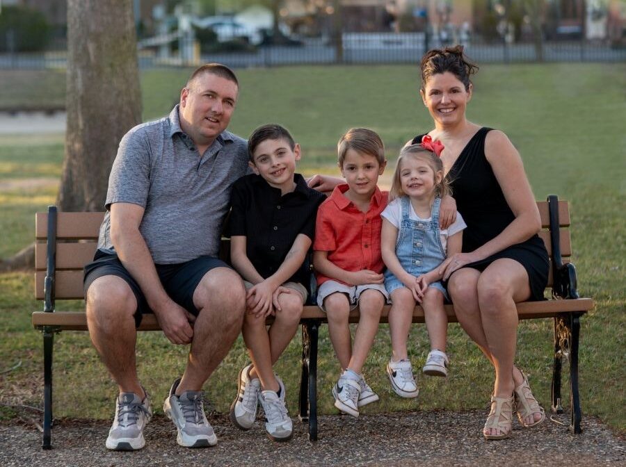 A family of five sits together on an outdoor bench. The dad sits on the left and the mom sits on the right; between them are two boys and a girl. Behind them is green grass and a tree.
