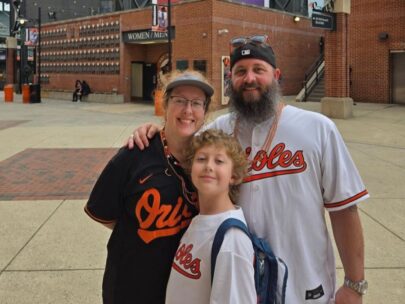 Photo shows three people: a woman, a man, and a boy. They are wearing Orioles baseball jerseys and standing in front of a stadium.
