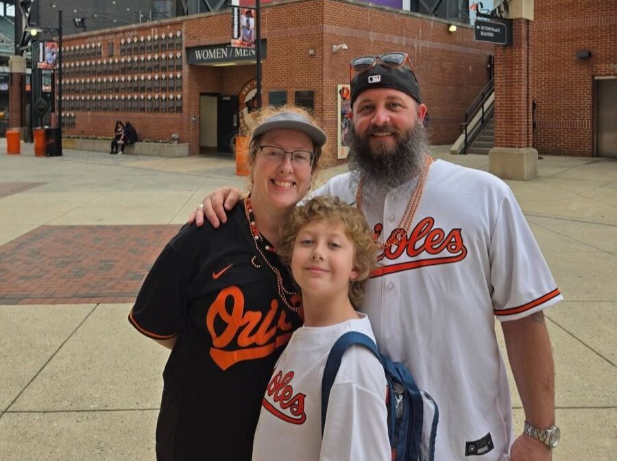 Photo shows three people: a woman, a man, and a boy. They are wearing Orioles baseball jerseys and standing in front of a stadium.