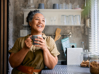 Stock photo shows a woman standing, leaning on a countertop, and holding a coffee mug. She appears to be looking out a window and smiling. Behind her are kitchen utensils and cups on shelves.