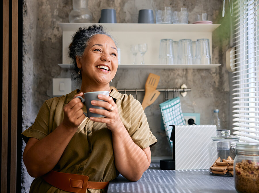 Stock photo shows a woman standing, leaning on a countertop, and holding a coffee mug. She appears to be looking out a window and smiling. Behind her are kitchen utensils and cups on shelves.