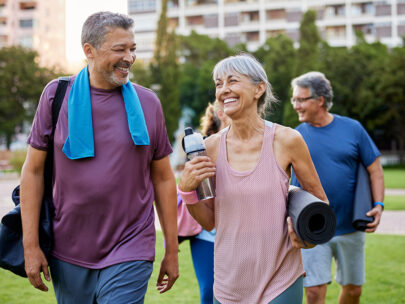 Stock photo shows an older man and woman walking. The woman is carrying a yoga mat and a water bottle, and the man is carrying a duffel bag and has a towel around his neck. They are outdoors, smiling and laughing with one another.
