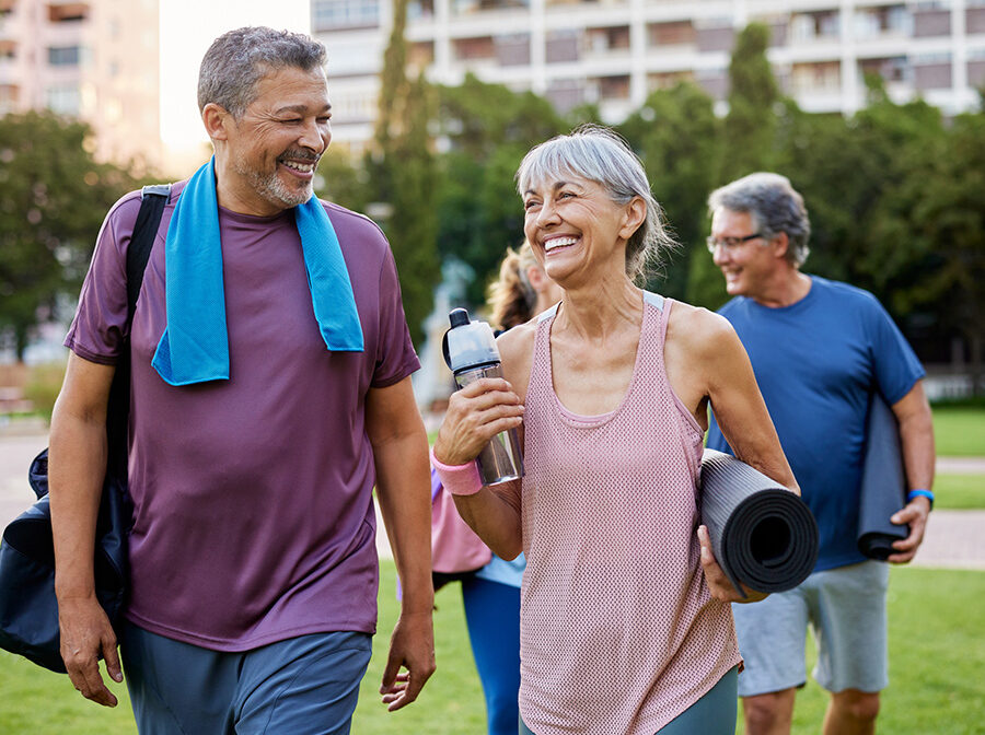 Stock photo shows an older man and woman walking. The woman is carrying a yoga mat and a water bottle, and the man is carrying a duffel bag and has a towel around his neck. They are outdoors, smiling and laughing with one another.