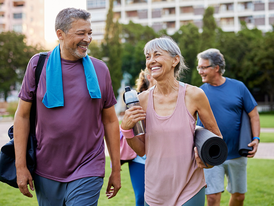 Stock photo shows an older man and woman walking. The woman is carrying a yoga mat and a water bottle, and the man is carrying a duffel bag and has a towel around his neck. They are outdoors, smiling and laughing with one another.