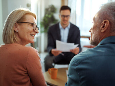 Stock photo shows an older man and woman looking at each other, smiling. Across the table from them in the background is a man holding papers.