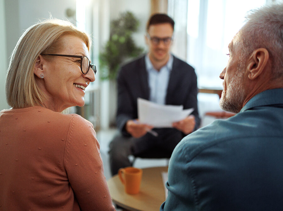 Stock photo shows an older man and woman looking at each other, smiling. Across the table from them in the background is a man holding papers.