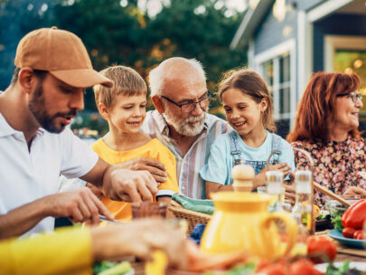 Stock photo shows an older man sitting outdoors with two kids on his lap, his arms around them. A young adult man is to their left, and an older woman is to their right. They are all seated at an outdoor table, on which are plates of food. Everyone looks happy and loving.