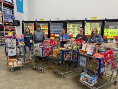 Photo shows five people standing in a store with four full shopping carts. In the carts are a wide variety of food, boxes of diapers, paper towels, and laundry detergent.
