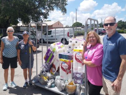 Four people stand around a cart holding donations. Among the donations are toilet paper, Kleenex, and other household items. The group is standing outside, where it is sunny.