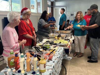 A line of people stand behind a long buffet table of food. One of the volunteers is wearing a Santa hat.
