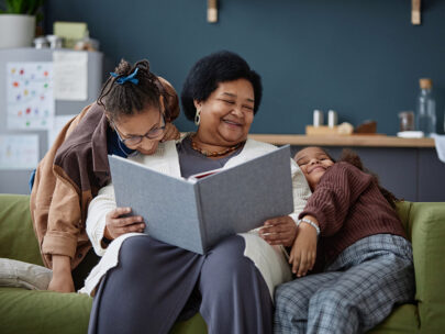 Stock photo shows an older woman sitting on a couch with two children. The children are leaning against her playfully, and she is smiling. She is holding a large open book.