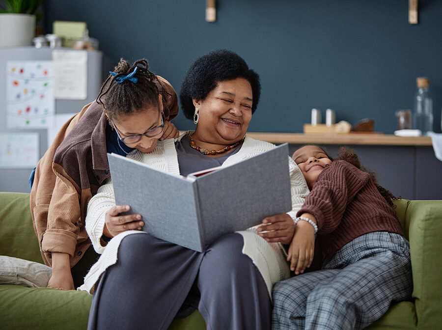 Stock photo shows an older woman sitting on a couch with two children. The children are leaning against her playfully, and she is smiling. She is holding a large open book.