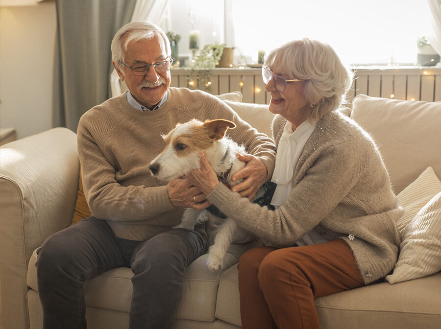 An older couple sit on a couch with a dog between them. Both are smiling as they pet the dog. There is a window behind them.