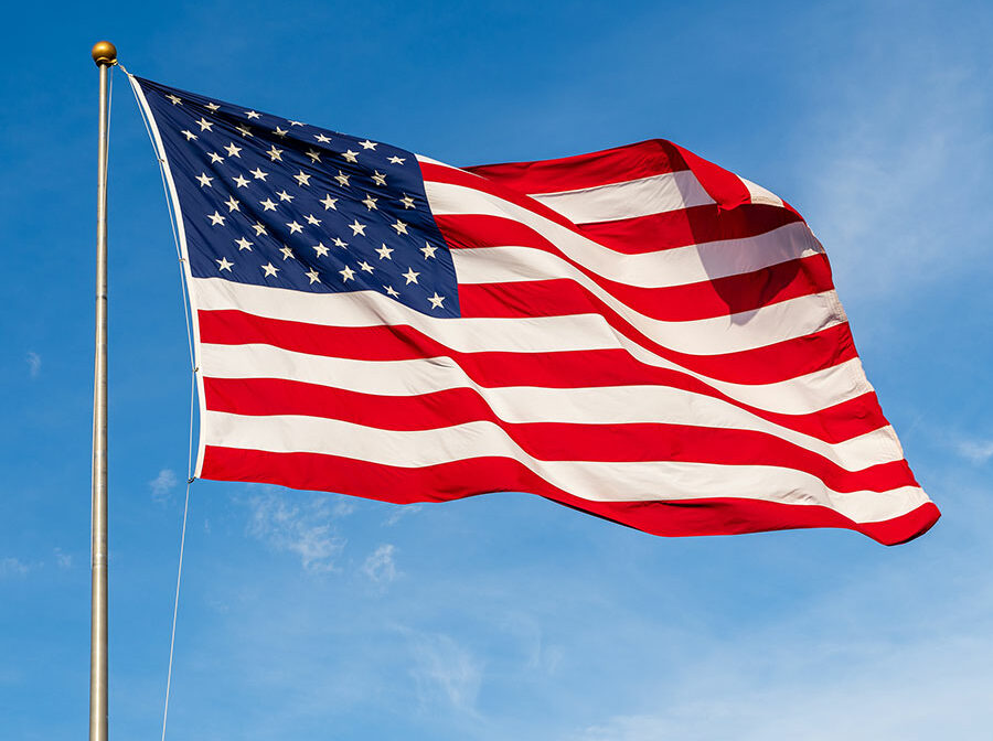 An American flag waves in the win at the top of a flagpole, with the blue sky as background.