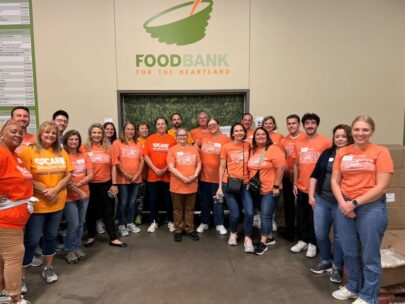 A group of 22 WoodmenLife associates stand together for a photo. They are all wearing orange WoodmenLife-branded shirts, and they are standing below a Food Bank for the Heartland logo on the wall.