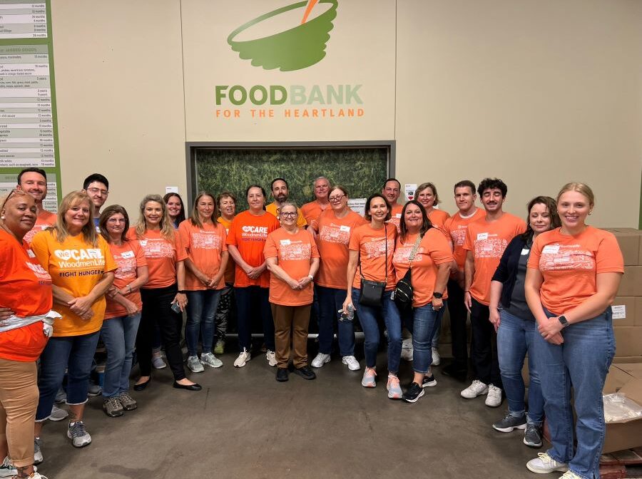 A group of 22 WoodmenLife associates stand together for a photo. They are all wearing orange WoodmenLife-branded shirts, and they are standing below a Food Bank for the Heartland logo on the wall.