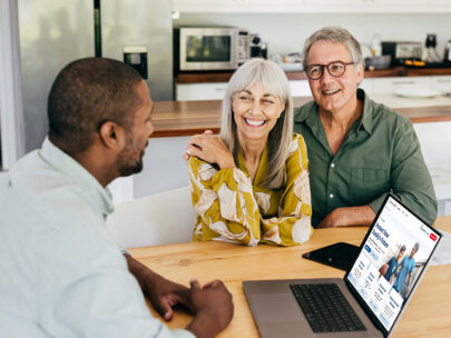 Stock photo shows an older couple sitting at a table with a professional. The professional has a laptop open in front of him. The couple are smiling.