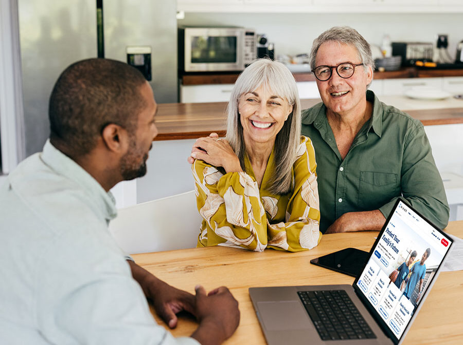 Stock photo shows an older couple sitting at a table with a professional. The professional has a laptop open in front of him. The couple are smiling.