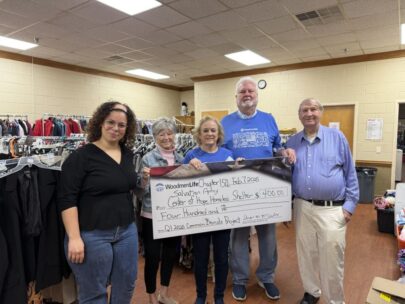 Five people stand together holding an oversized check. The check is made out to Center of Hope Homeless Shelter in the amount of $400. Behind the people are racks of donated clothing.