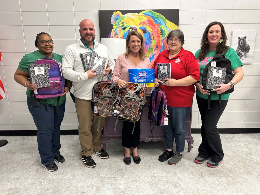 Five people stand in a school hallway. They are holding donations for the school, including backpacks, notebooks and Clorox wipes.