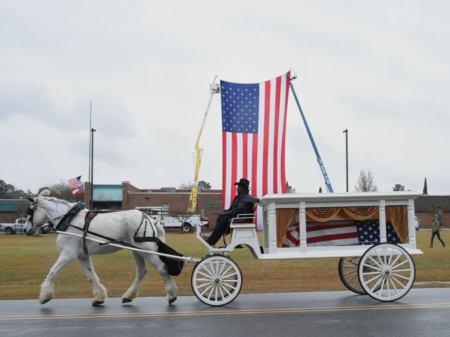 A flag-draped casket is transported on a horse-drawn carriage. Behind that is a 30-foot-by-60-foot American flag, held up in the air vertically by two cranes.