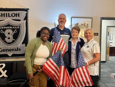 Four people — three women and one man — stand together. They are all holding 12-by-18-inch American flags on flag poles.