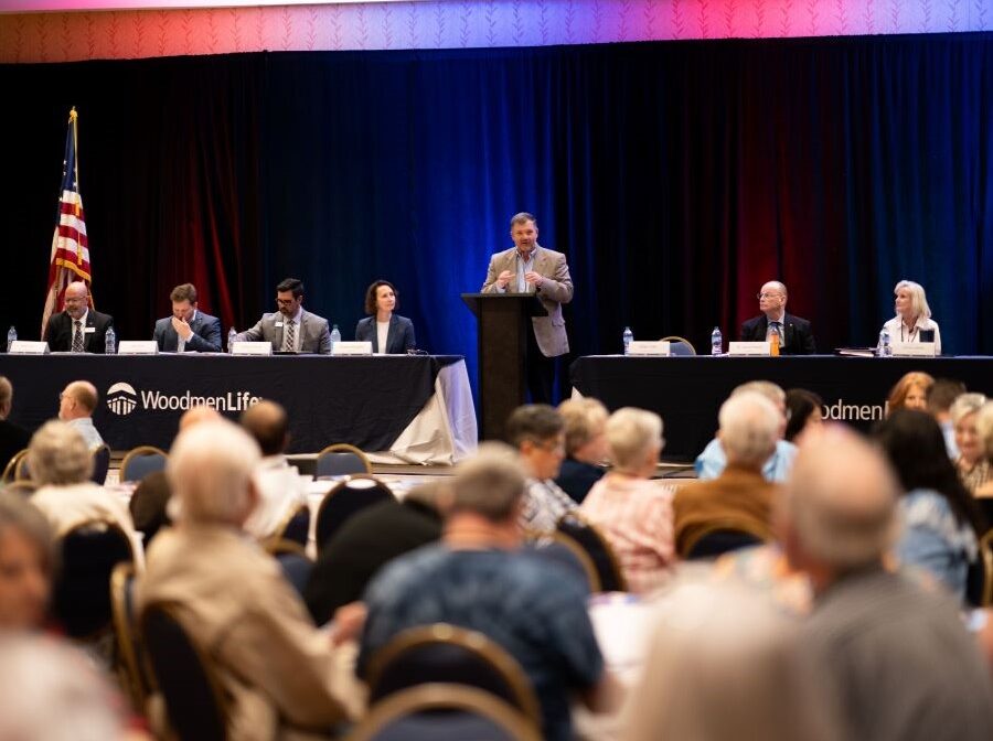 Photo is looking toward a stage. There is someone speaking at a podium in the center of the stage, and long tables on either side of the podium at which people are seated. In the foreground of the photo are the backs of people's heads as they listen.