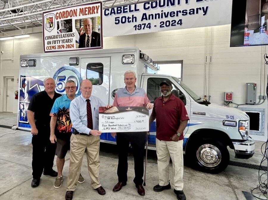 Five men pose for a photo around an oversized WoodmenLife-branded check. Behind them is an ambulance. Behind them and hanging from the ceiling is a banner that says "Gordon Merry, Congratulations on fifty years! CCEMS 1975-2024." A second banner reads "Cabell County EMS, 50th Anniversary, 1974-2024."