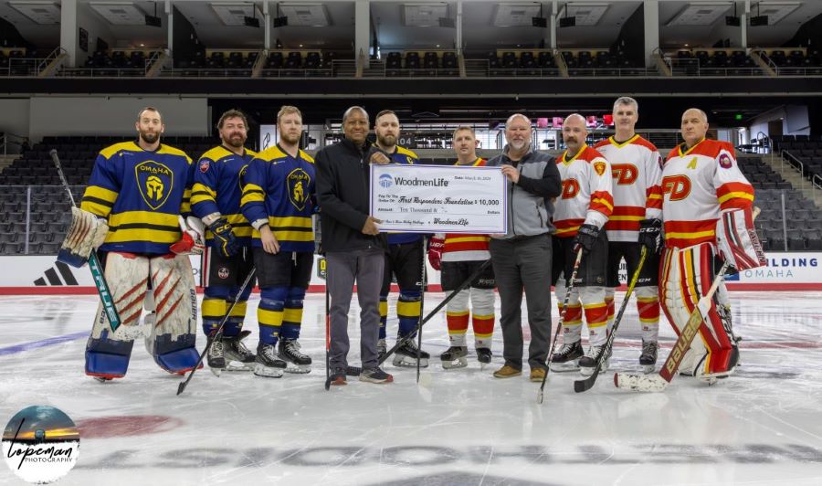 Photo shows a total of 10 men posing for a photo at an ice rink. Eight of the men are in hockey uniforms; four of them are wearing blue and yellow jerseys representing the police and four are wearing red, white and yellow jerseys representing the firefighters. At the center of the group, two men in plain clothes hold an oversized check showing WoodmenLife's $10,000 donation to the First Responders Foundation.