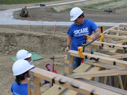 Three people are shown at a house being constructed. One person is on a ladder, and he is looking down at the two people standing on the ground. All three people are wearing blue T-shirts that say "WeCare WoodmenLife", and they're each wearing white hard hats.
