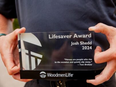 This photo is a close-up of a WoodmenLife Heart & Heritage Award. It is a glass award, about 6 inches wide and 4 inches tall. The award pictured says "Lifesaver Award, Josh Shedd, 2024" plus a quote at the bottom that says "Heroes are people who rise to the occasion and quietly slip away. - Tom Brokaw." The WoodmenLife logo is at the bottom of the award.