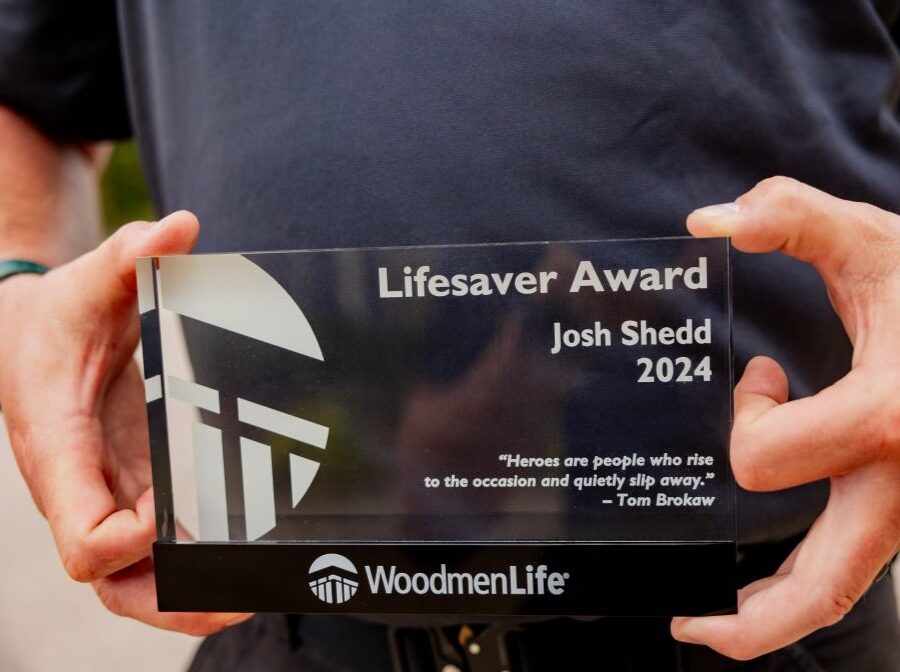 This photo is a close-up of a WoodmenLife Heart & Heritage Award. It is a glass award, about 6 inches wide and 4 inches tall. The award pictured says "Lifesaver Award, Josh Shedd, 2024" plus a quote at the bottom that says "Heroes are people who rise to the occasion and quietly slip away. - Tom Brokaw." The WoodmenLife logo is at the bottom of the award.