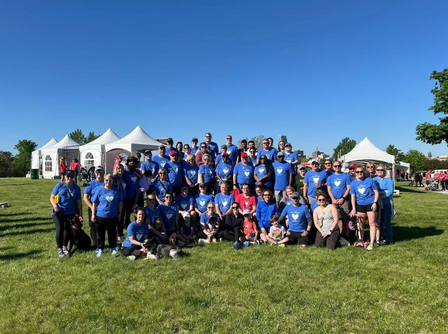 A large group of people pose for a photo. Most people are wearing matching blue T-shirts. The group is outside on grass; behind them are the tops of white tents.