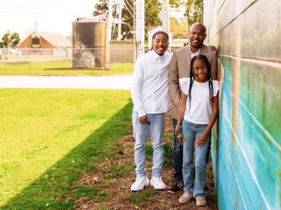 A man poses for the photo with two children, a son and a daughter. They are leaning against a wall with colorful artwork. Behind them is green grass with sunlight shining on it.
